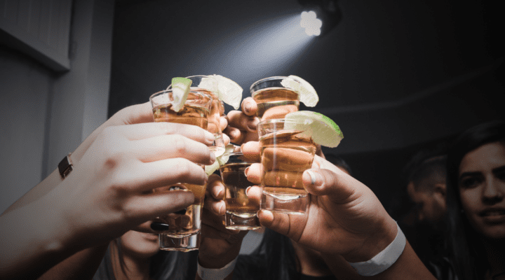 A group of women raise their lime-rimmed glasses, filled with a light-colored tequila, and cheers at center frame